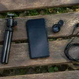 Чорний смартфон, міні-штатив, кліп-об’єктив і навушники на дерев’яній лавці