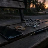 Темна дерев’яна лавка зі смартфоном, навушниками, монетами і ручкою на ній