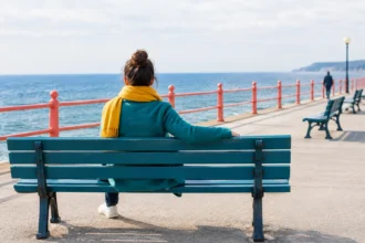 Жінка в пальті й жовтому шарфі сидить на лавці біля моря